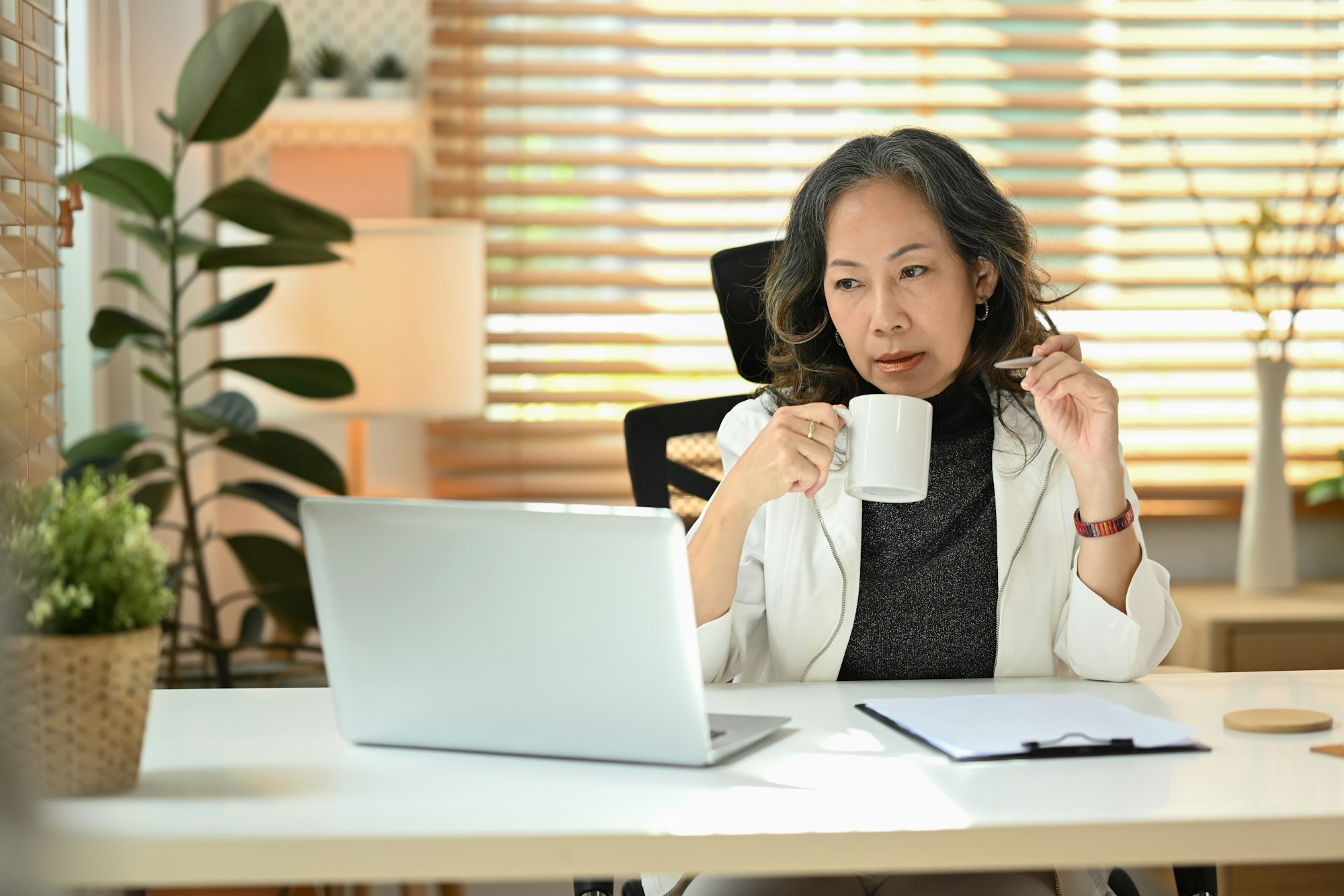 Senior businesswoman drinking coffee and watching online webinar during working online .